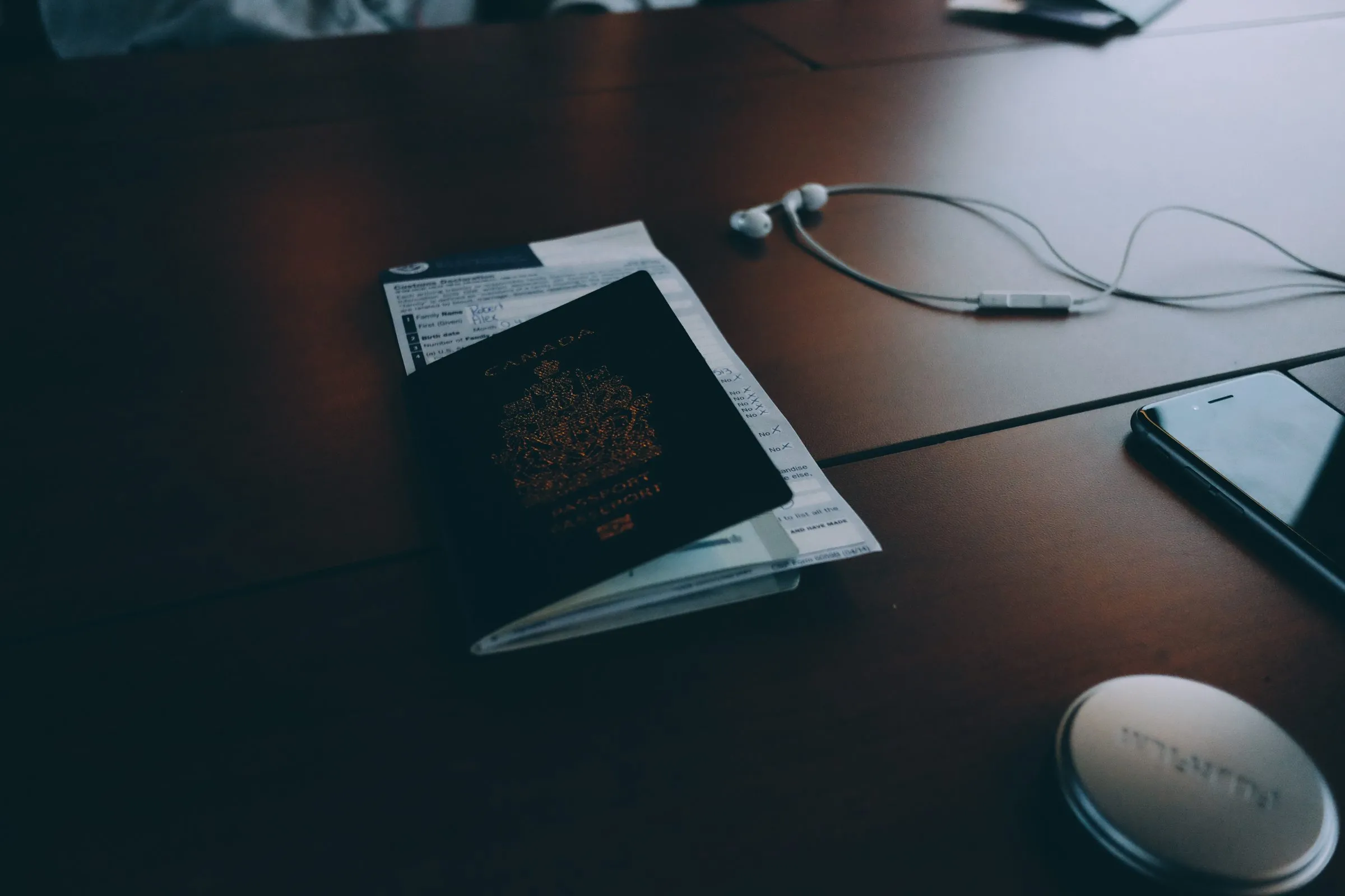 A closed Canadian passport lying on a dark wooden desk next to a customs form, earphones and a phone