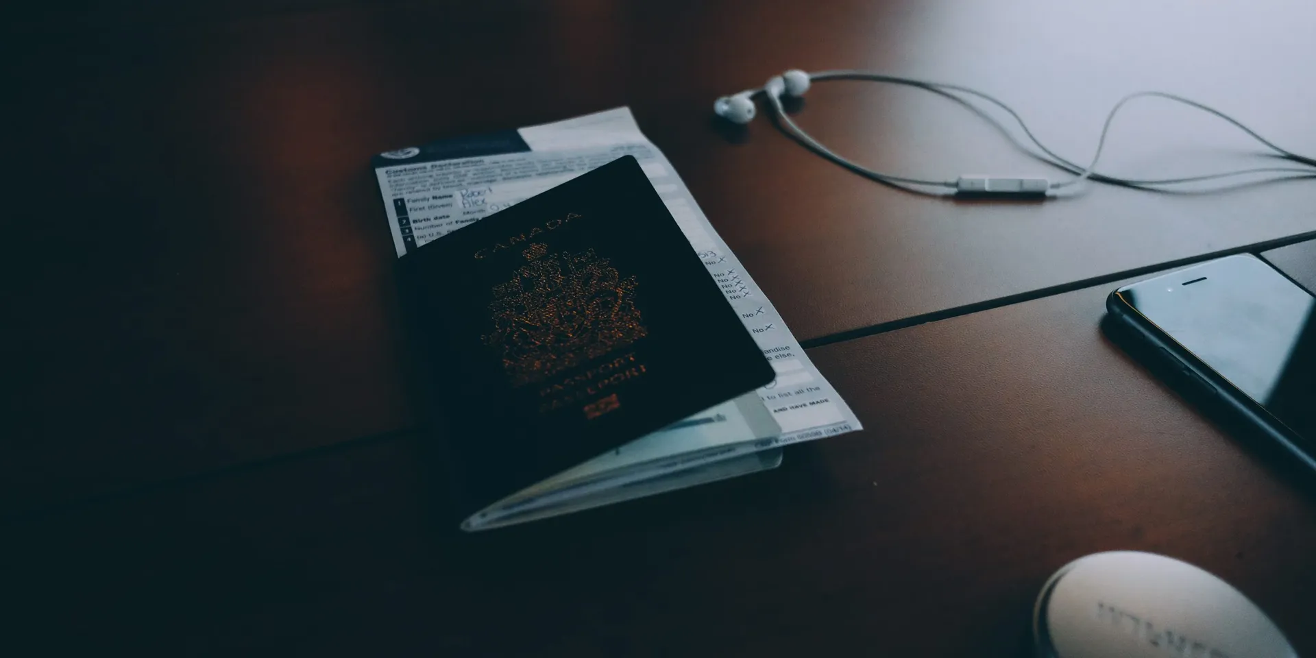 A closed Canadian passport lying on a dark wooden desk next to a customs form, earphones and a phone